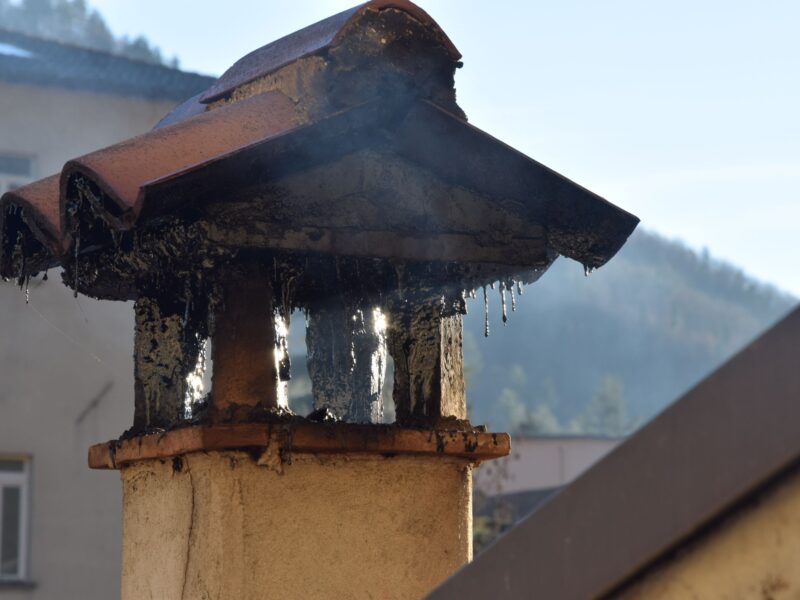 Close-up of a burnt metal roof gutter atop a beige column, with melted tar dripping down and a blurred building in the background.