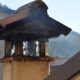 Close-up of a burnt metal roof gutter atop a beige column, with melted tar dripping down and a blurred building in the background.