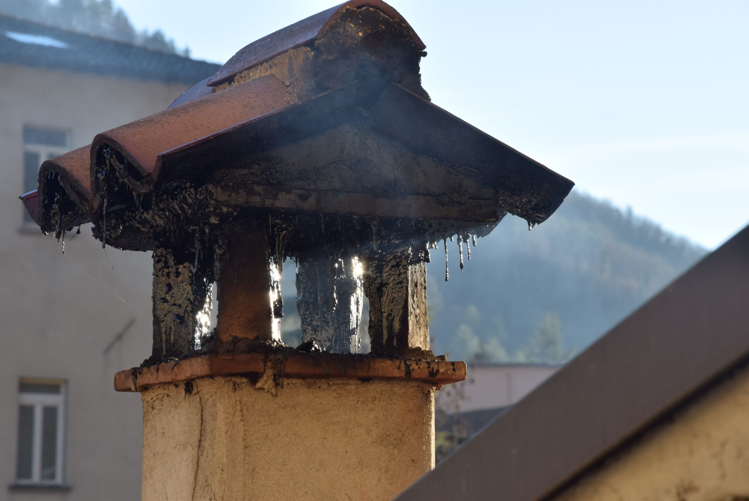Close-up of a burnt metal roof gutter atop a beige column, with melted tar dripping down and a blurred building in the background.